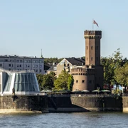 Malakoff Tower Am Ufer des Rheins in Köln erhebt sich der historische Malakoffturm, flankiert von modernen Gebäuden und umgeben von Bäumen, die in der Nachmittagssonne leuchten.The historic Malakoff Tower stands on the banks of the Rhine in Cologne, flanked by modern buildings and surrounded by trees that glow in the afternoon sun.