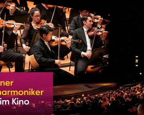 Ein Orchester spielt auf einer Bühne in einem halbdunklen, gut besetzten Konzertsaal.