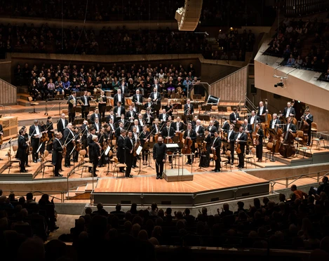 Das Bild zeigt die Berliner Philharmoniker in einem modernen Saal, umgeben von einem aufmerksamen Publikum.