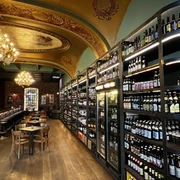 Ein langer Gang führt bis an das andere Ende des Lokals. In einem großen Wandregal rechts stehen viele verschiedene Flaschen. Links vom Gang stehen Holztische und Holzstühle sowie Barhocker, die an der langen Theke links stehen. Die Decke ist sehr hoch und gewölbt und mit goldenen Verzierungen versehen.