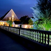 Wilsmann Apartments Hövelhof bei Nacht, beleuchtet mit umgebenden Pflanzen und einem dekorativen Zaun.
