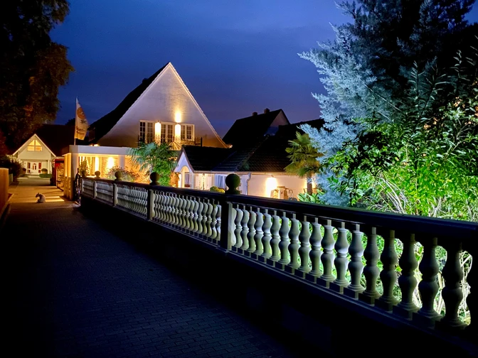 Wilsmann Apartments Hövelhof bei Nacht, beleuchtet mit umgebenden Pflanzen und einem dekorativen Zaun.