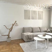 Helles Wohnzimmer einer Ferienwohnung mit grauem Sofa, weißen Tischen und großen Fenstern.