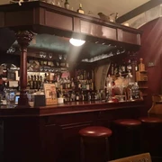 Kleine Theke aus dunklem Holz, in der sich Regale mit Gläsern und verschiedenen Flaschen befinden. Vor der Theke stehen mehrere Barhocker.