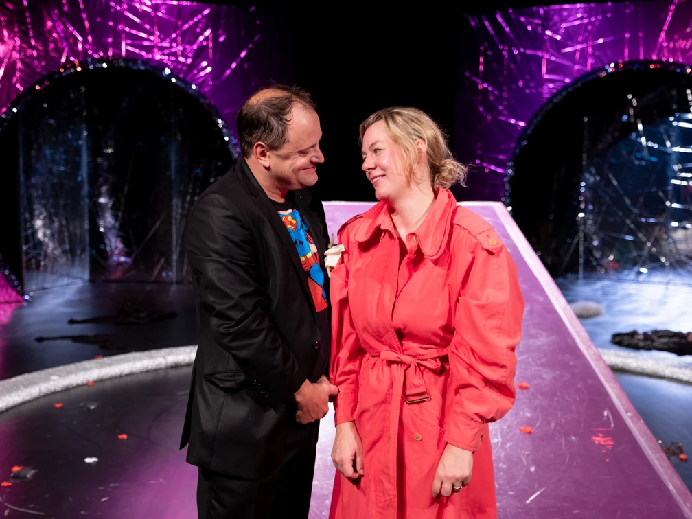 Ein Mann und eine Frau stehen lächelnd auf einer Bühne mit farbenfrohem Hintergrund.