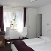 Doppelzimmer mit zwei weißen Betten, Schreibtisch, zwei Stühlen und einem Fenster mit Vorhängen.