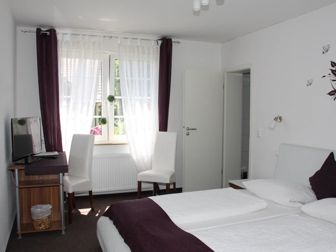 Doppelzimmer mit zwei weißen Betten, Schreibtisch, zwei Stühlen und einem Fenster mit Vorhängen.