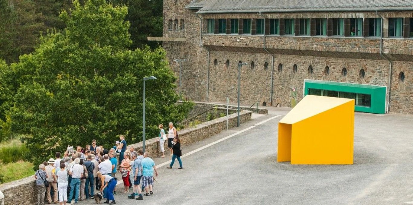 Personengruppe steht vor modernem, gelbem Kunstwerk, historische Burg Vogelsang im Hintergrund.