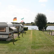 Ruhiger Campingplatz in Wassernähe mit Wohnwagen und Fahnen im Sommer, umgeben von grüner Natur.