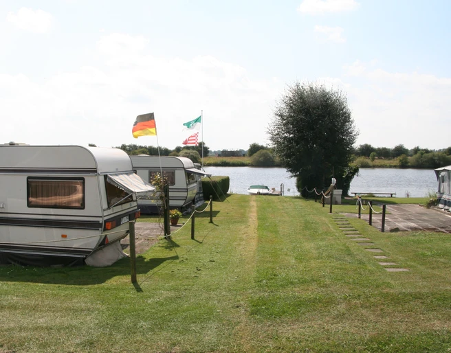 Ruhiger Campingplatz in Wassernähe mit Wohnwagen und Fahnen im Sommer, umgeben von grüner Natur.