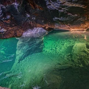 Besucherbergwerk "Marie Louise Stolln" Unterwasseraufnahme einer Höhle mit grünlich schimmerndem Wasser, Felsen und einer Leiter am Boden, beleuchtet von warmem Licht.
