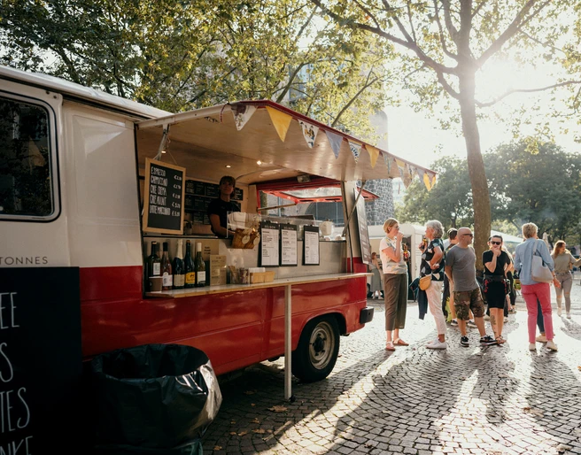 meet & eat Cologne Ein farbenfroher Foodtruck auf einem sonnigen Platz, umgeben von Menschen, die in entspannter Atmosphäre Essen genießen.A colorful food truck in a sunny square, surrounded by people enjoying food in a relaxed atmosphere.