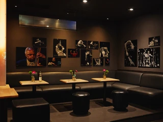Stadtgarten Ein gemütlicher, dunkel gehaltener Raum mit schwarzen Ledersitzmöglichkeiten und Jazz-Fotografien an den Wänden.A cozy, dark room with black leather seating and jazz photographs on the walls.