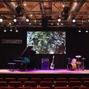 Stadtgarten Eine Bühne mit Flügel, Gitarre, Schlagzeug und "Stadtgarten"-Schild im Hintergrund.A stage with a grand piano, guitar, drums and a "Stadtgarten" sign in the background.