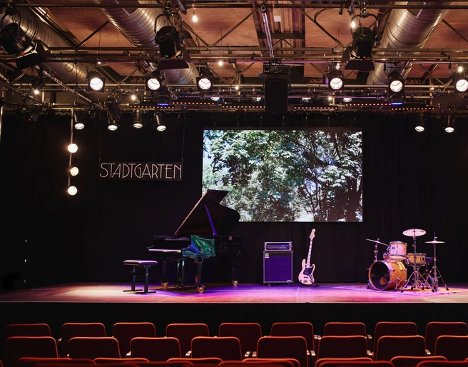 Stadtgarten Eine Bühne mit Flügel, Gitarre, Schlagzeug und "Stadtgarten"-Schild im Hintergrund.A stage with a grand piano, guitar, drums and a "Stadtgarten" sign in the background.