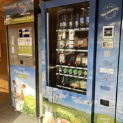 Milchautomat Bichlerhof, Farchant im kleinen Holzhäusl steht linkerhand ein Milchautomat mit Flaschen, rechts davon zwei Automaten mit Yogurt, Wurst und Fleischauswahl, vakumiert. in der Mitte findet sich der Bezahlautomat