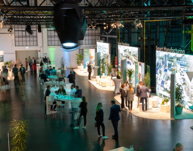 Fujitsu Smart Solutions In einem großen, modernen Veranstaltungsraum sind verschiedene Gruppen von Menschen versammelt. Helle Lichter und große Bildschirme, die verschiedene Themen rund um "Smart Manufacturing" präsentieren, dominieren die Szenerie. Die Atmosphäre ist belebt, mit Gesprächen und Networking an Stehtischen.Various groups of people are gathered in a large, modern event room. Bright lights and large screens presenting various topics related to smart manufacturing dominate the scene. The atmosphere is lively, with conversations and networking at bar tables.