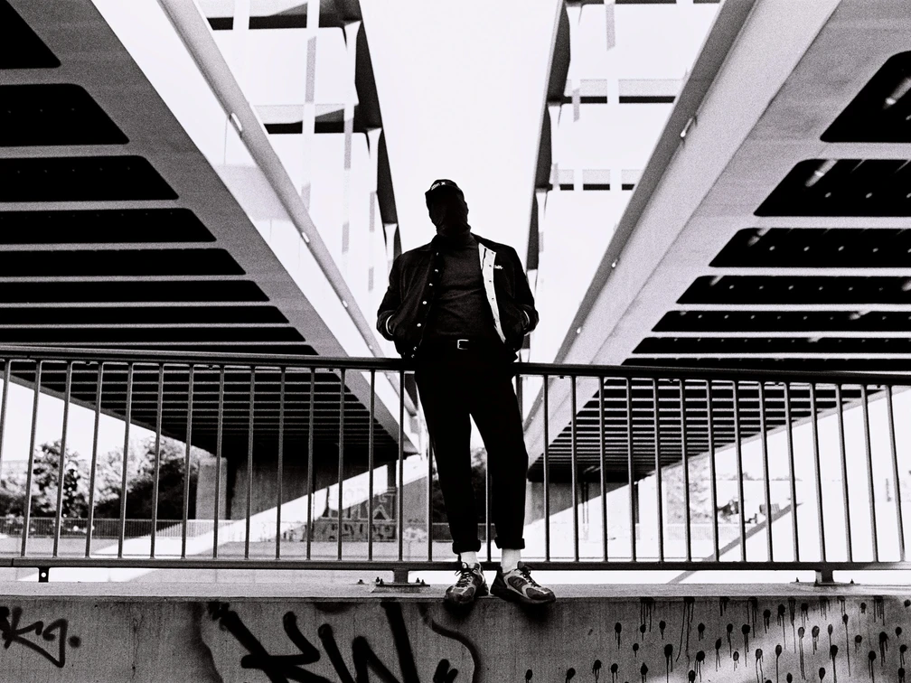 PoebelMC_Pressefotos2021_FotocreditMarshlCeronPalomino_5-scaled.jpg Ein Mann in urbaner Kulisse steht vor Geländer unter Brückenstruktur, kontrastreich in Schwarzweiß.A man in an urban setting stands in front of railings under a bridge structure, rich in contrast in black and white.Een man in een stedelijke omgeving staat voor leuningen onder een brugstructuur, contrastrijk in zwart-wit.