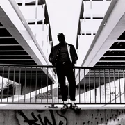 PoebelMC_Pressefotos2021_FotocreditMarshlCeronPalomino_5-scaled.jpg Ein Mann in urbaner Kulisse steht vor Geländer unter Brückenstruktur, kontrastreich in Schwarzweiß.A man in an urban setting stands in front of railings under a bridge structure, rich in contrast in black and white.Een man in een stedelijke omgeving staat voor leuningen onder een brugstructuur, contrastrijk in zwart-wit.
