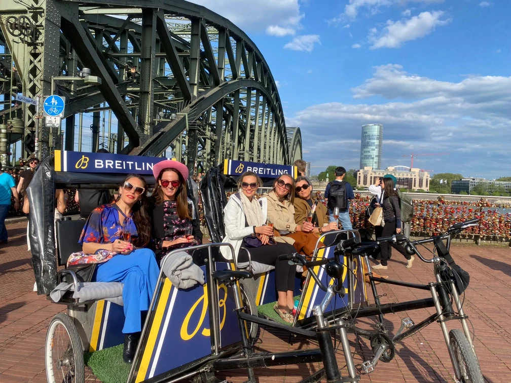 Rikolonia Champagnerfahrt.jpg Fünf Frauen sitzen in einer Fahrrad-Rikscha vor einer Brücke mit Vorhängeschlössern und genießen den Ausblick.</p>Five women sit in a bicycle rickshaw in front of a padlocked bridge and enjoy the view.</p> <p