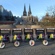Rikolonia_Event Branding.jpg Die farbenfrohen Fahrradrikschas stehen vor dem eindrucksvollen Kölner Dom in der Abenddämmerung.The colorful bicycle rickshaws stand in front of the impressive Cologne Cathedral at dusk.