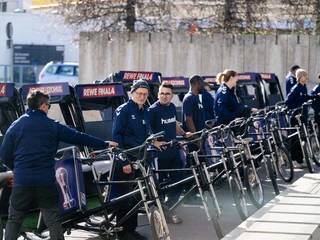 Rikolonia_Event VIP Shuttle.jpeg Menschen in blauer Kleidung mit Fahrradrikschas aufgereiht in urbaner Umgebung.</p>People in blue clothing lined up with bicycle rickshaws in an urban environment.</p> <p