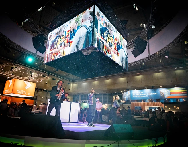 Frauenthal-Expo-Wien-PLUS-Messeband-Entertainment-DJplus-04.jpg Musiker auf einer Messebühne, große Bildschirme zeigen lebhafte Szenen, umgeben von Publikum.</p>Musicians on an exhibition stage, large screens show lively scenes, surrounded by an audience.</p> <p