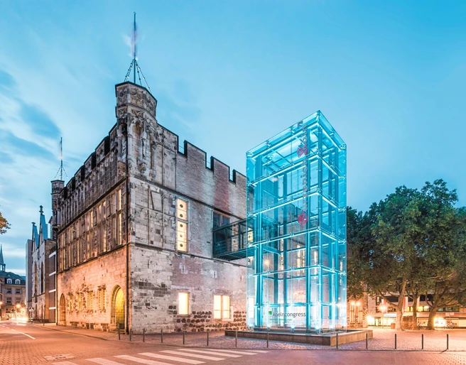 Gürzenich Köln, Außenansicht Altes Rathaus mit modernem gläsernen Bau im Abendlicht, umgeben von Bäumen und beleuchteter Straße.</p>Old town hall with modern glass building in the evening light, surrounded by trees and illuminated street.</p> <p
