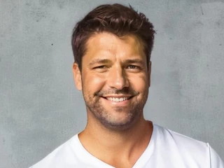 Foto.jpg Ein lächelnder Mann mit braunem Haar im weißen T-Shirt vor einer grauen, strukturierten Wand.