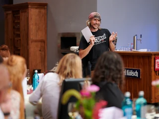 Cologne Graffiti. Der Coach Ein Mann hält fröhlich ein Blatt Papier und spricht vor einer Gruppe in einem Raum mit rustikaler Bar.A man cheerfully holds a piece of paper and speaks to a group in a room with a rustic bar.