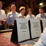 Hopfen & Malz. Genuss-Quiz Tisch <p>Personen diskutieren an einem Tisch über Quizfragen in einem stimmungsvoll beleuchteten Raum.</p>