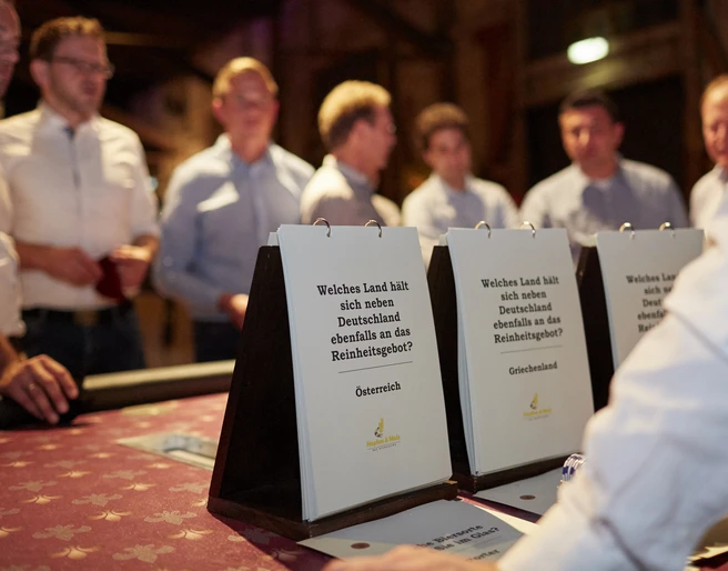 Hopfen & Malz. Genuss-Quiz Tisch <p>Personen diskutieren an einem Tisch über Quizfragen in einem stimmungsvoll beleuchteten Raum.</p>