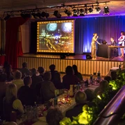 Köln ist ein Gefühl. Die Comedy Musical Dinner Show Musical Dinner Show: Auf der Bühne stehen drei Schauspieler neben einem Bild mit der Kölner Skyline. Zahlreiche Gäste sitzen an langen Banketttischen und schauen die ShowMusical dinner show: Three actors stand on stage next to a picture of the Cologne skyline. Numerous guests sit at long banquet tables and watch the show