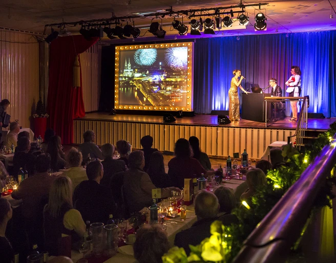Köln ist ein Gefühl. Die Comedy Musical Dinner Show Musical Dinner Show: Auf der Bühne stehen drei Schauspieler neben einem Bild mit der Kölner Skyline. Zahlreiche Gäste sitzen an langen Banketttischen und schauen die ShowMusical dinner show: Three actors stand on stage next to a picture of the Cologne skyline. Numerous guests sit at long banquet tables and watch the show
