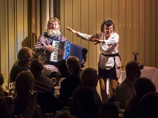 Köln ist ein Gefühl. Die Comedy Musical Dinner Show. Servicefachkraft Akkordeonspieler in Begleitung einer fröhlichen Frau, die für ein Publikum auftritt.Accordion player accompanied by a cheerful woman performing for an audience.