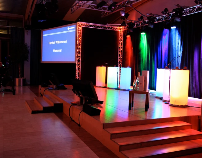 WeQuiz. Das Quizevent. Bühne Beleuchtete Bühne mit mehrfarbigen Scheinwerfern, Podium und Projektorbildschirm "Herzlich Willkommen".</p>Illuminated stage with multicolored spotlights, podium and "Welcome" projector screen.</p> <p