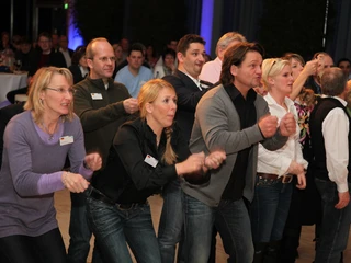 WeQuiz. Das Quizevent. Aktivrunde Menschen in Freizeitkleidung tanzen synchron in einer Gruppe in einem Veranstaltungsraum.</p>People in casual clothes dance synchronously in a group in an event room.</p> <p