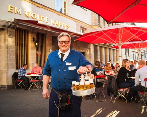 Früh Em Golde Kappes Auf dem Bild ist ein Kellner zu sehen, der eine blaue Jacke trägt und einen Kranz mit Biergläsern in der Hand hält. Er lächelt freundlich in die Kamera. Im Hintergrund sitzen Gäste an Tischen mit roten Sonnenschirmen vor einem Gebäude, auf dem "Em Golde Kappes" steht. Die Szene vermittelt eine gesellige Stimmung in einer Außengastronomie.The picture shows a waiter wearing a blue jacket and holding a wreath of beer glasses. He smiles friendly into the camera. In the background, guests are sitting at tables with red parasols in front of a building that reads "Em Golde Kappes". The scene conveys a convivial atmosphere in an outdoor restaurant.