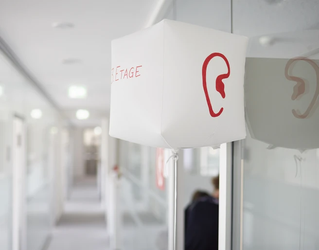 SINNfonie. Ein Parcours durch die Welt de Sinne Ein Flur mit moderner Beleuchtung und einem roten Ohrsymbol auf einer weißen Leuchte.A hallway with modern lighting and a red ear symbol on a white luminaire.