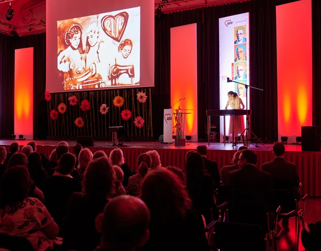 AWO 100-jähriges Jubiläum.jpg Konzertsaal mit rotem Licht, Musikerin am Keyboard, große Projektion von Kunstwerken an der Wand.</p>Concert hall with red light, musician at the keyboard, large projection of artworks on the wall.</p> <p