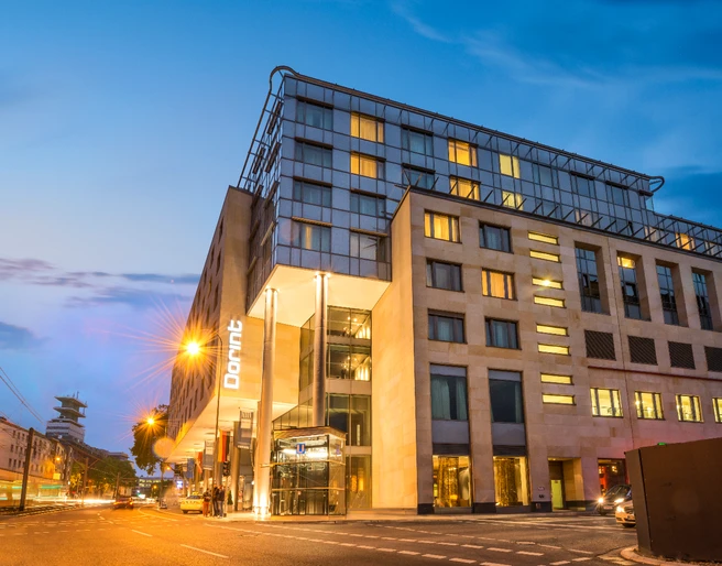 Dorint Hotel am Heumarkt  Modernes Hotel mit beleuchteter Fassade bei Dämmerung, Straßenbahn fährt vorbei.</p>Modern hotel with illuminated façade at dusk, streetcar passes by</p>.
