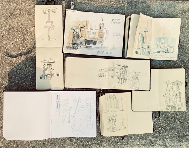 Urban Sketchers Mehrere geöffnete Skizzenbücher auf dem Boden zeigen unterschiedlich detaillierte urbane Zeichnungen von Straßenszenen mit Menschen und Architektur.