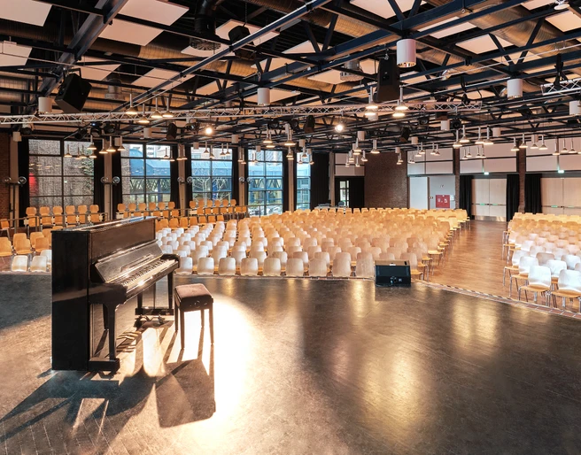 Großer Saal Großer, leerer Veranstaltungssaal mit Holzstühlen und einem schwarzen Klavier auf der Bühne.</p>Large, empty event hall with wooden chairs and a black piano on the stage.</p> <p
