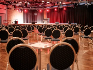 Bistrobestuhlung Großer Saal Bestuhlter Veranstaltungssaal mit Bühne, rotem Licht und Klavier, bereit für eine Aufführung.</p>Seated event hall with stage, red light and piano, ready for a performance.