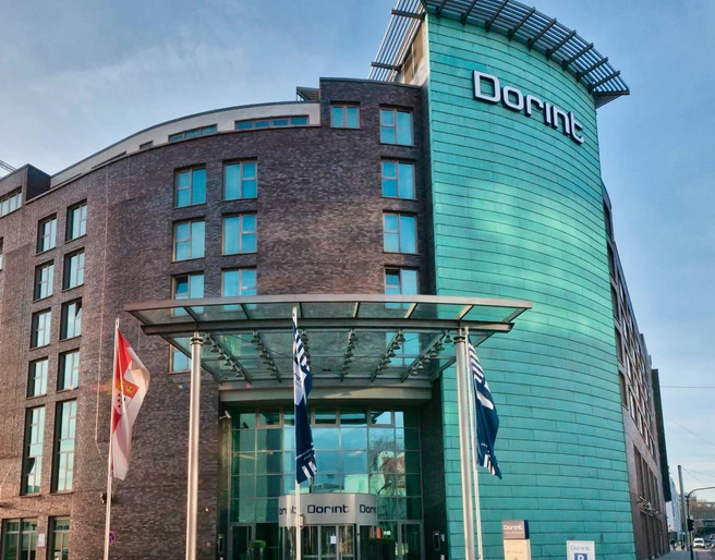 Dorint Hotel an der Messe Köln  Modernes Hotelgebäude mit Glasfassade und zwei Flaggen im Vordergrund.</p>Modern hotel building with glass façade and two flags in the foreground.</p> <p
