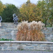 Laura Ford "A & E Monkey" (2012) seit 2017 in Eschborn Kunstinstallation mit 3 Affen, die auf einer Mauer sitzen. Sie haben unterschiedliche Körperteile verbunden. Vor der Muer ist ein mittelhohes Federgras gepflanzt, davor eine Sitzbank.