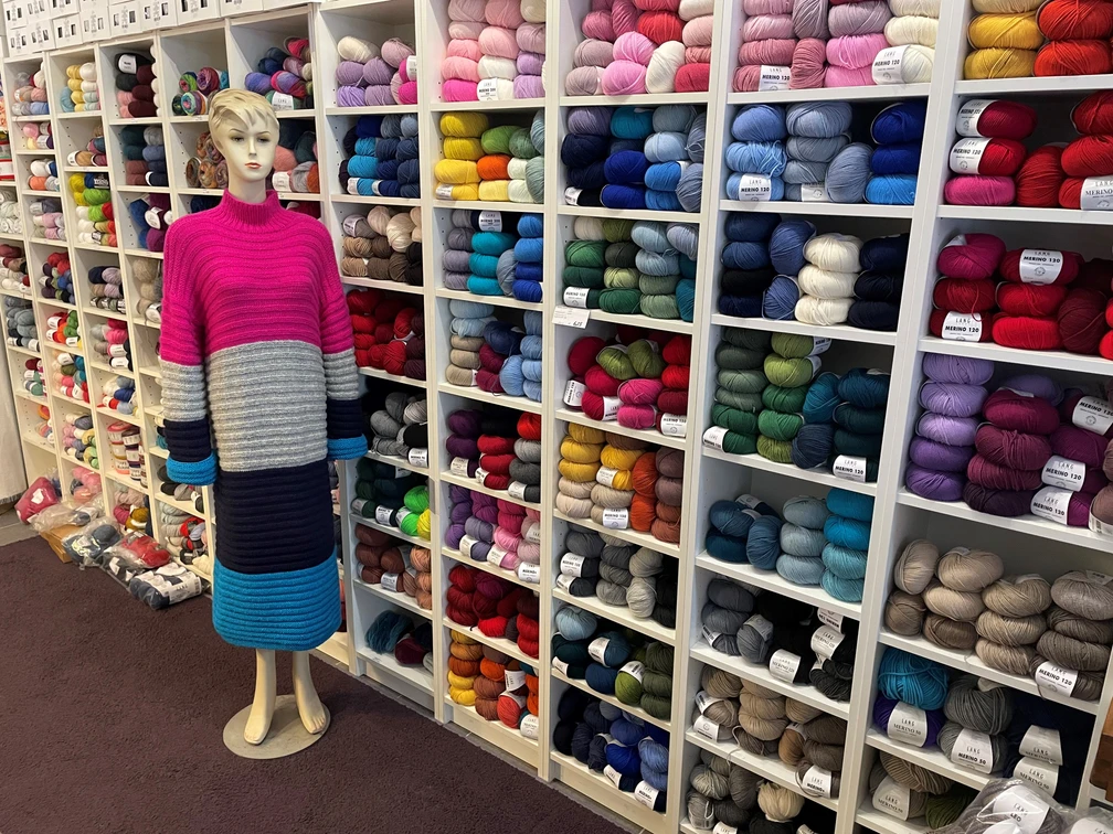 maris.masche1.jpg Eine Schaufensterpuppe im langen, gestreiften Kleid steht vor einem Regal voller bunter Wollknäuel.