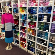 maris.masche1.jpg Eine Schaufensterpuppe im langen, gestreiften Kleid steht vor einem Regal voller bunter Wollknäuel.