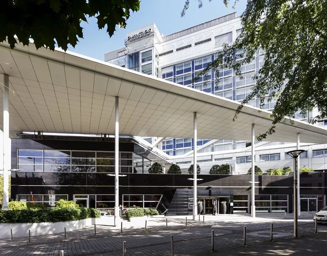 Pullman Cologne 5366 Das Bild zeigt die moderne Frontansicht des Pullman Cologne Hotels mit seiner markanten Architektur und großen Fenstern. Vor dem gepflegten Gebäude erstreckt sich eine überdachte Vorfahrt mit schlanken Säulen, die einen einladenden Eingangsbereich schaffen.The picture shows the modern front view of the Pullman Cologne Hotel with its striking architecture and large windows. A covered driveway with slender columns extends in front of the well-kept building, creating an inviting entrance area.