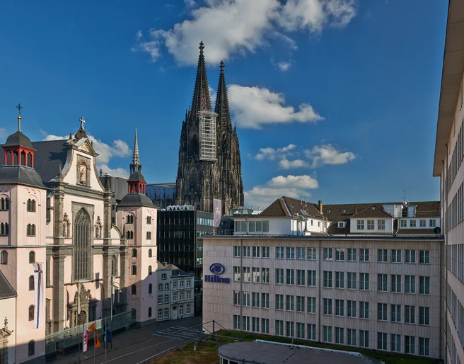 Hilton Cologne Kölner Dom mit steinernen Türmen und umliegenden Gebäuden bei blauem Himmel und leichter Bewölkung.Cologne Cathedral with stone towers and surrounding buildings under a blue sky and light cloud cover.